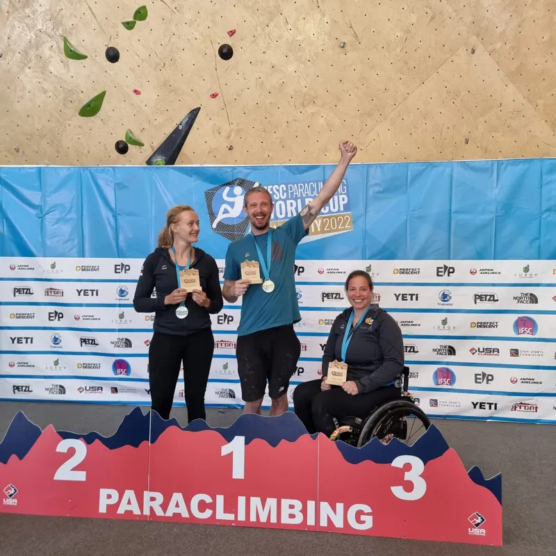 Drie glimlachende paraklimmers staan met hun medailles op het podium, waarop de plaatsen 1, 2 en 3 zijn aangegeven. Op de derde plaats staat een vrouw in een rolstoel. Op de achtergrond zijn een klimwand en een blauw evenementbanner te zien.